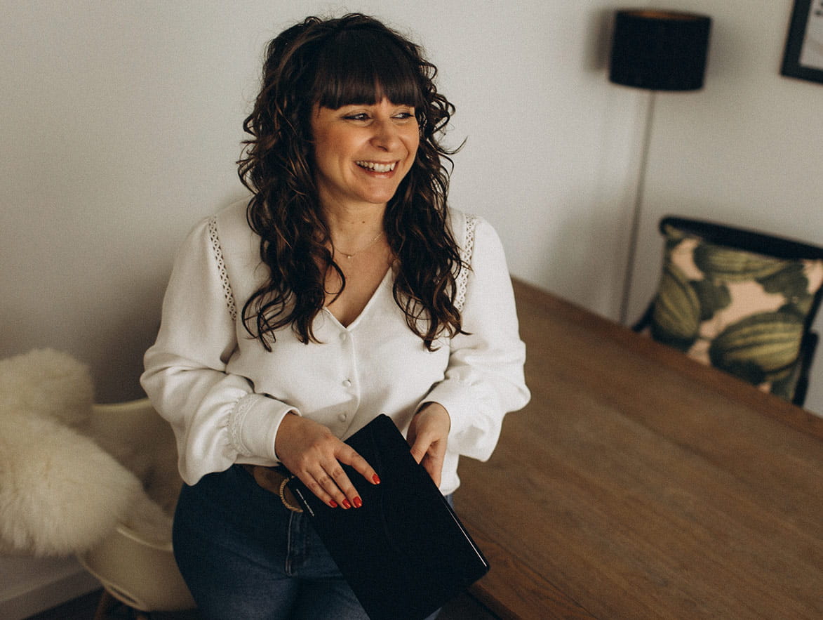 Photo de Julie Venturini souriant, assise contre son bureau, tenant un cahier